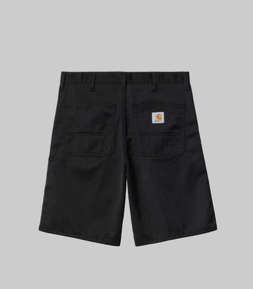 Black Carhartt work shorts shown from the back, featuring two large patch pockets and the signature Carhartt logo patch on the right pocket, set against a solid gray background.