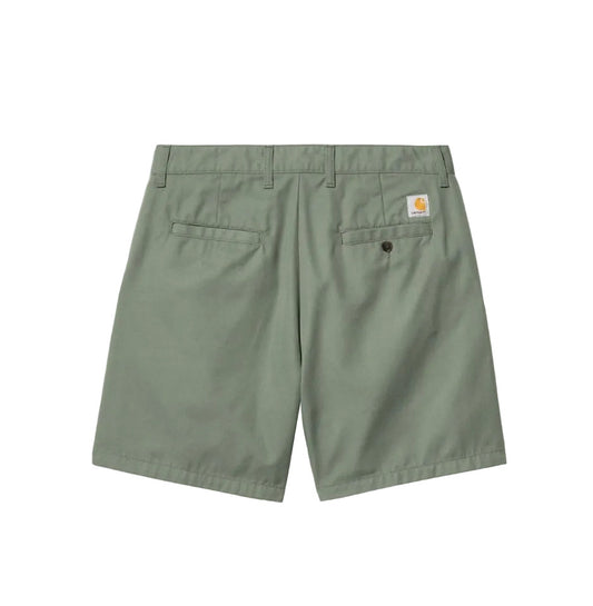 A pair of sage green Carhartt shorts shown from the back, featuring two back pockets and a small signature Carhartt logo patch on the right pocket, set against a solid gray background.