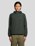 A man wearing a dark green hooded jacket with a small yellow Lyle logo on the chest, paired with olive green pants, against a solid gray background.