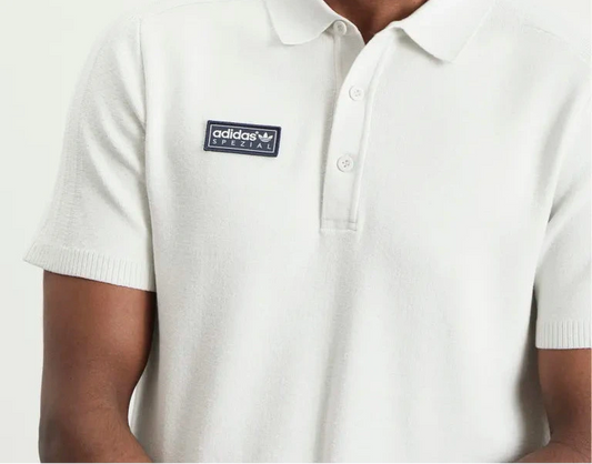 A close-up of a man wearing a white short-sleeve polo shirt with a black Adidas Originals logo patch on the left chest, set against a plain light background.