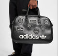A person wearing black Adidas pants and black loafers holds a black Adidas duffel bag covered in white and multicolor patch designs against a light gray studio background.