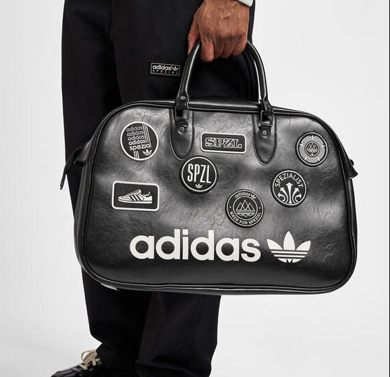 A person wearing black Adidas pants and black loafers holds a black Adidas duffel bag covered in white and multicolor patch designs against a light gray studio background.