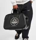 A person wearing a white zip-up hoodie and black sweatpants holds a black duffel bag with a white mountain logo and the word 'SPEZIAL' on a light gray studio background.