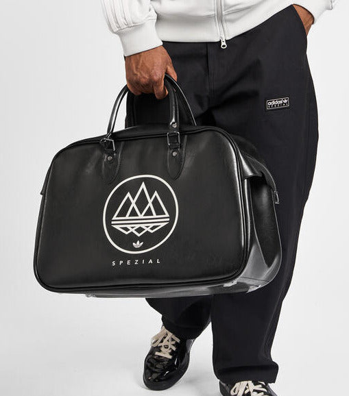 A person wearing a white zip-up hoodie and black sweatpants holds a black duffel bag with a white mountain logo and the word 'SPEZIAL' on a light gray studio background.
