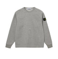 A light gray heather crewneck sweatshirt with raglan sleeves, featuring the black and yellow Stone Island compass patch on the left sleeve, displayed against a neutral light gray background.