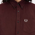 A close-up of a model wearing a dark burgundy corduroy button-down shirt by Fred Perry, featuring the signature embroidered laurel wreath logo on the chest pocket.