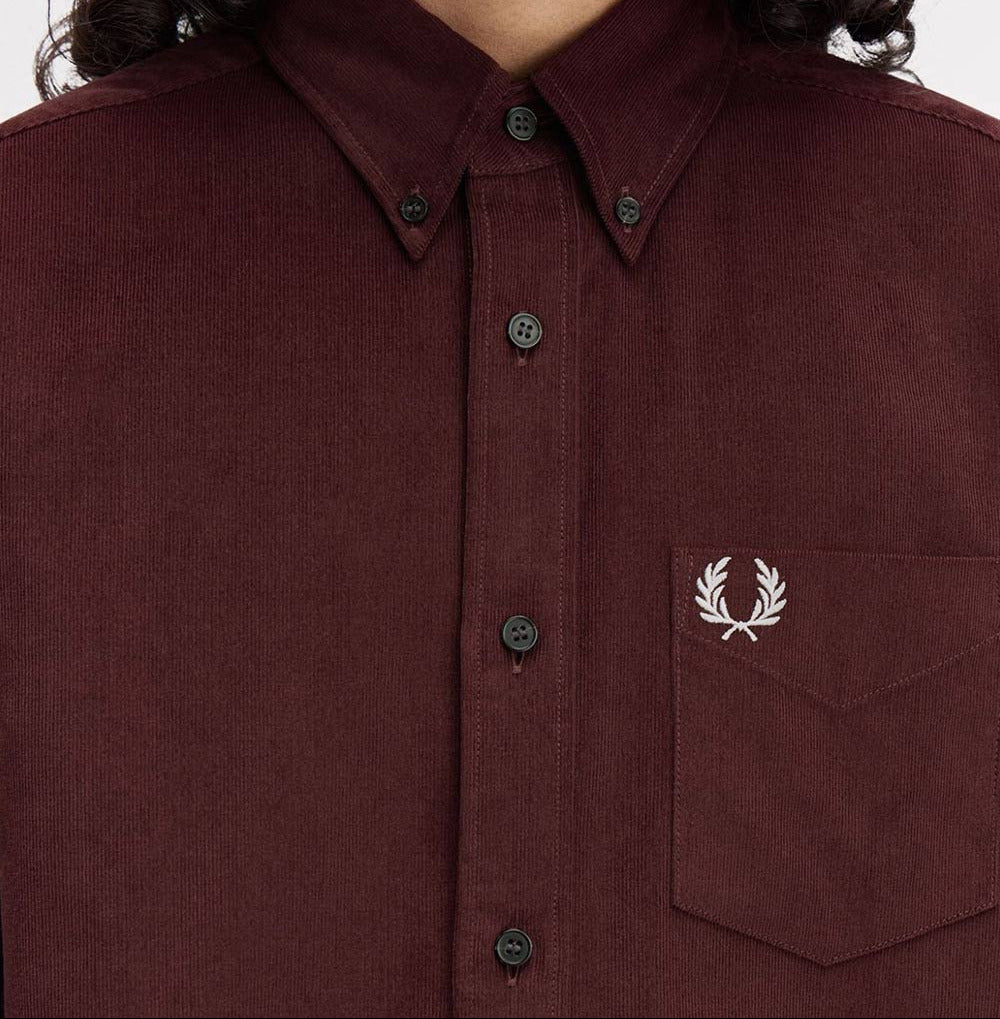 A close-up of a model wearing a dark burgundy corduroy button-down shirt by Fred Perry, featuring the signature embroidered laurel wreath logo on the chest pocket.
