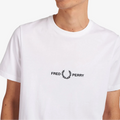 A smiling young man with short blond hair wearing a white crew-neck t-shirt with the black 'FRED PERRY' logo and laurel wreath printed on the chest, set against a plain white background.