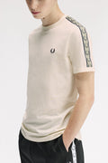A man wearing a cream-colored short-sleeve t-shirt by Fred Perry, featuring a black embroidered laurel wreath logo on the left chest and a green-and-white patterned stripe down the right sleeve. He is also wearing black track pants with a matching white-and-green patterned side stripe.