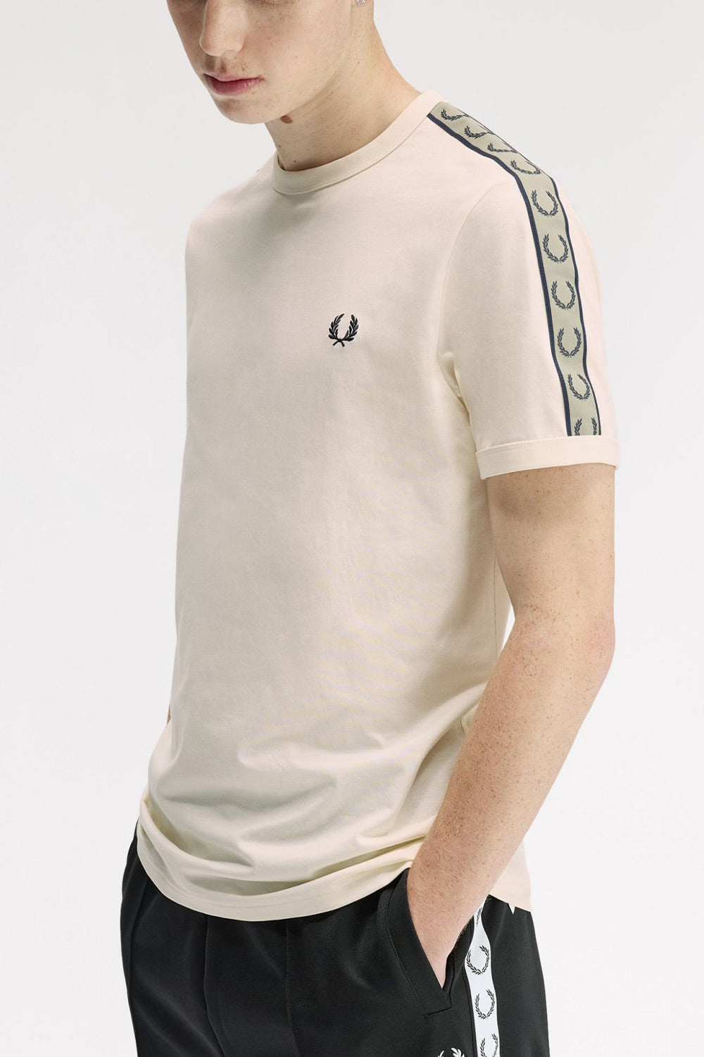 A man wearing a cream-colored short-sleeve t-shirt by Fred Perry, featuring a black embroidered laurel wreath logo on the left chest and a green-and-white patterned stripe down the right sleeve. He is also wearing black track pants with a matching white-and-green patterned side stripe.