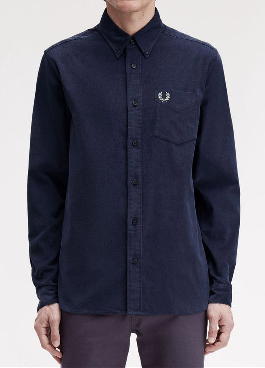 A man wearing a dark navy long-sleeve button-up shirt by Fred Perry, featuring the signature embroidered laurel wreath logo on the left chest pocket, set against a plain light gray background.