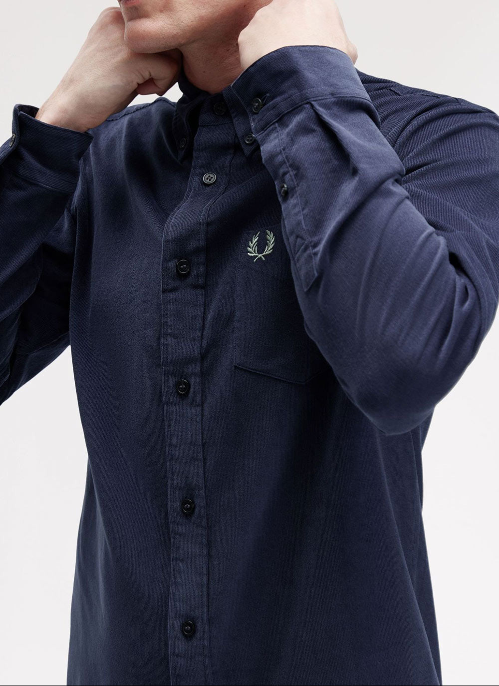 A close-up of a man wearing a navy blue long-sleeve button-up shirt by Fred Perry, featuring the signature embroidered laurel wreath logo on the left chest.