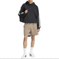 A model wearing a black Adidas hoodie with white stripes on the sleeves, beige shorts, white socks, and white sneakers, standing against a plain white background.