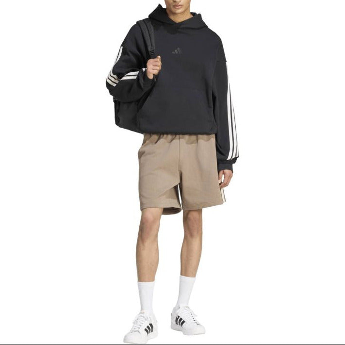 A model wearing a black Adidas hoodie with white stripes on the sleeves, beige shorts, white socks, and white sneakers, standing against a plain white background.