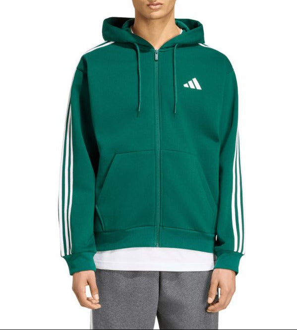 A man wearing a green Adidas full-zip hoodie with white stripes on the sleeves and a white logo on the chest, paired with gray sweatpants, against a plain white background.